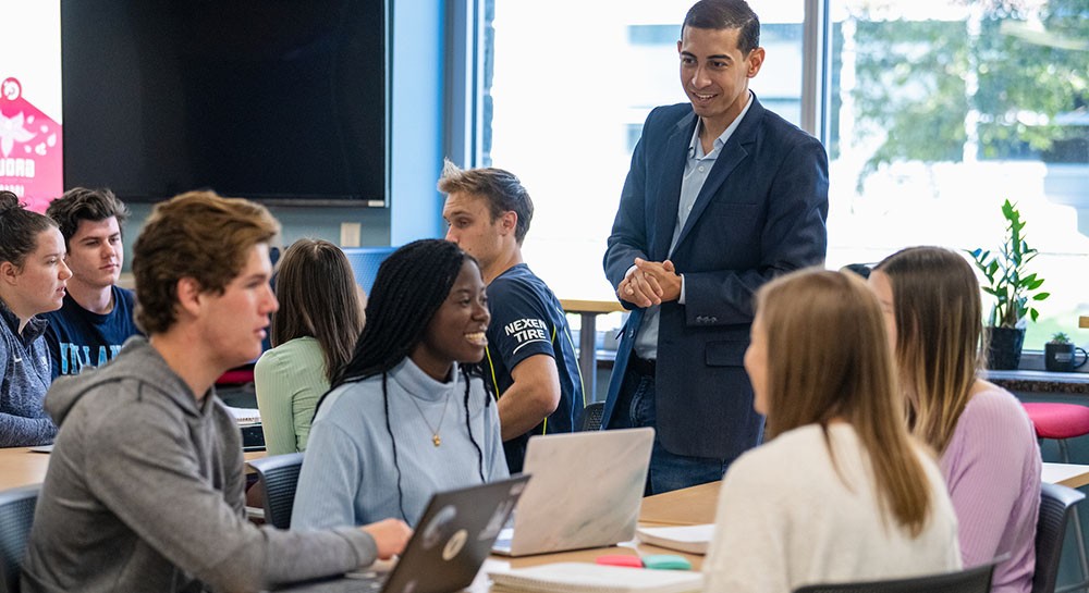 Faculty member standing and talking to group of students seated around a table
