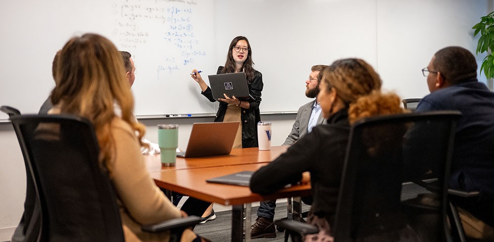 student presenting data at whiteboard to group seated at table