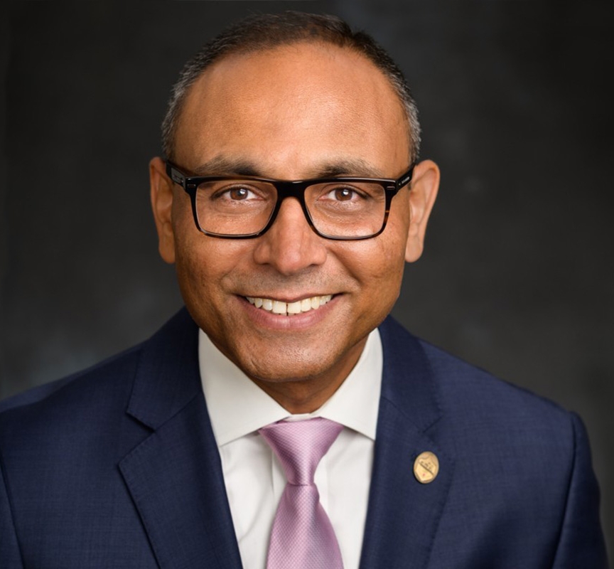 Headshot of Tej Patel smiling directly at the camera wearing a blue suit on a gray backdrop.