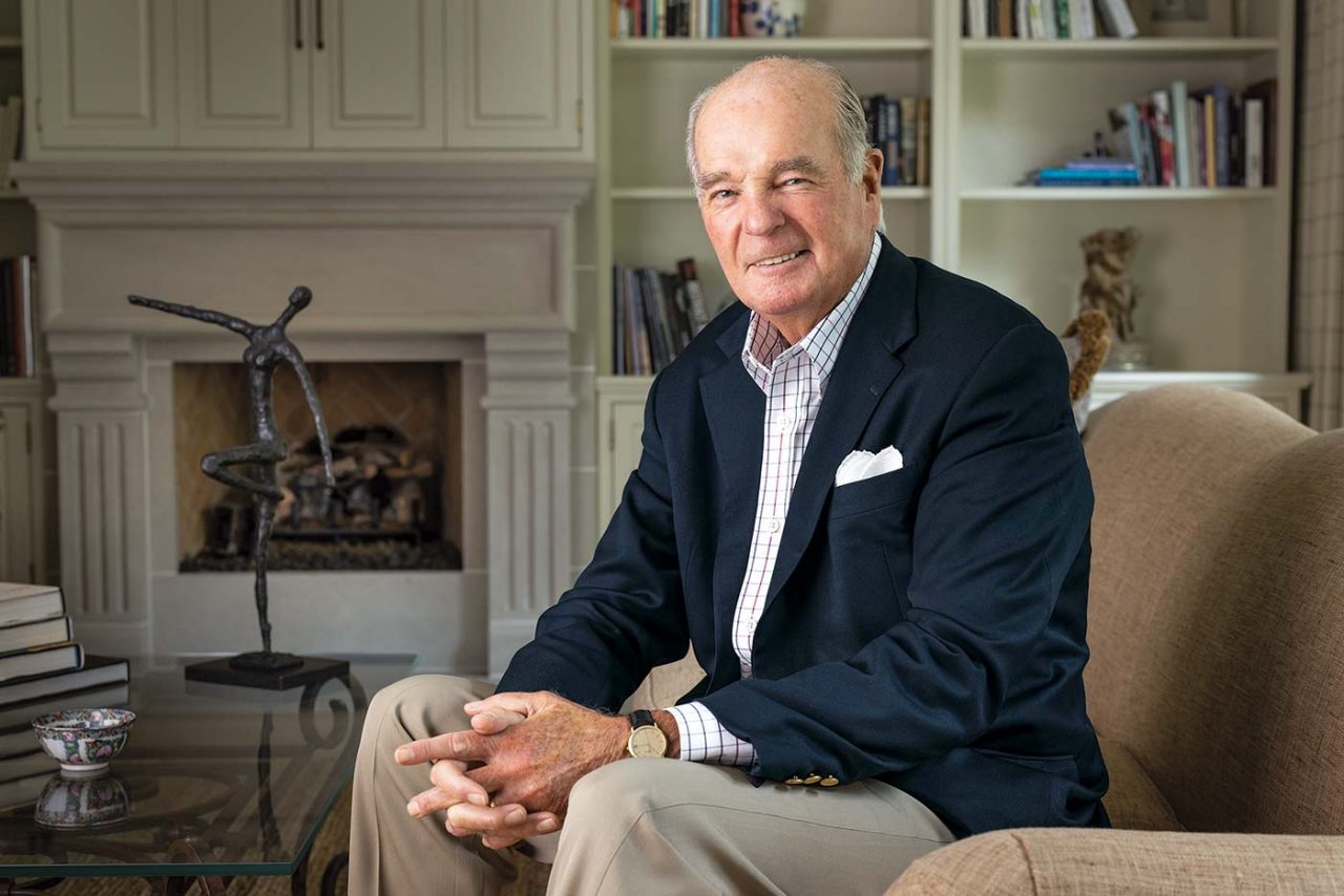 Martin McGuinn hands folded sitting in a chair against the backdrop of bookshelves and a fireplace